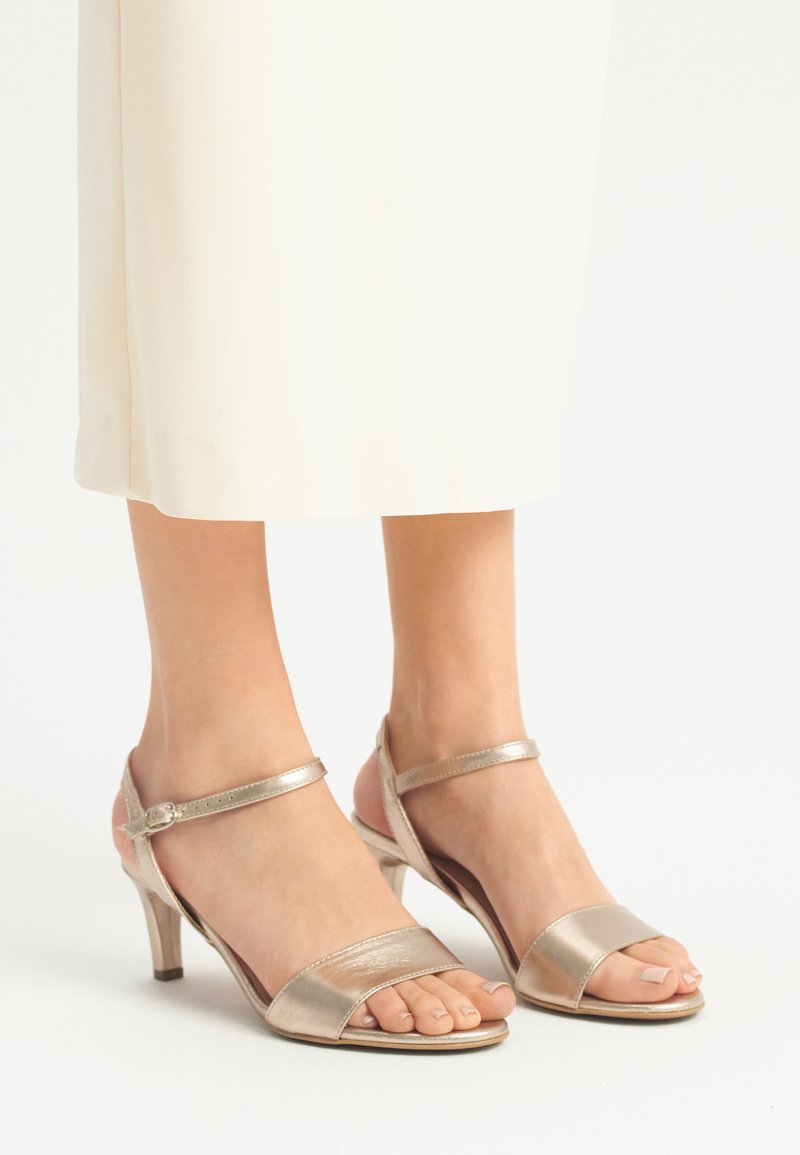 Metallisch glänzende goldene High-Heelsandalen mit offener Zehenpartie und Knöchelriemen, die eine glänzende Oberfläche und ein zugespitztes Absatzdesign aufweisen.