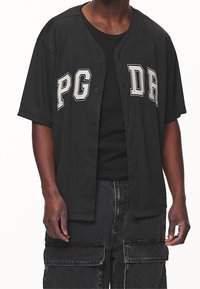 Uomo che indossa un maglione nero con bottoni e la scritta bianca "PG DR" sopra una maglietta a coste nera e pantaloni cargo neri.