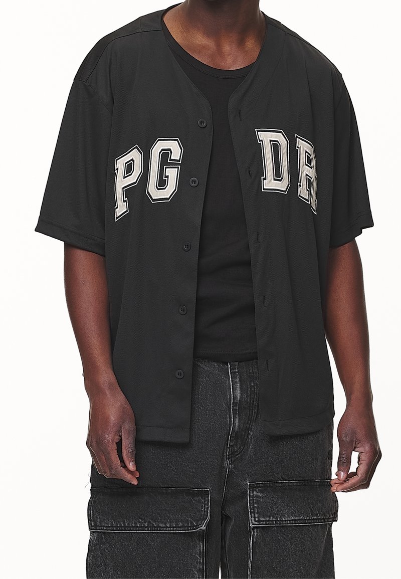 Uomo che indossa un maglione nero con bottoni e la scritta bianca "PG DR" sopra una maglietta a coste nera e pantaloni cargo neri.