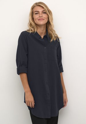 Plavuša s valovitom kosom u tamnoplavoj košulji s dugim rukavima i otkrivenim rukavima, stoji ispred jednobojne bijele pozadine.