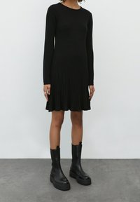 Robe noire à manches longues avec des côtes, dotée d'un ourlet évasé, associée à des bottes noires à semelle épaisse montantes jusqu'au genou. Texture lisse et silhouette ajustée.