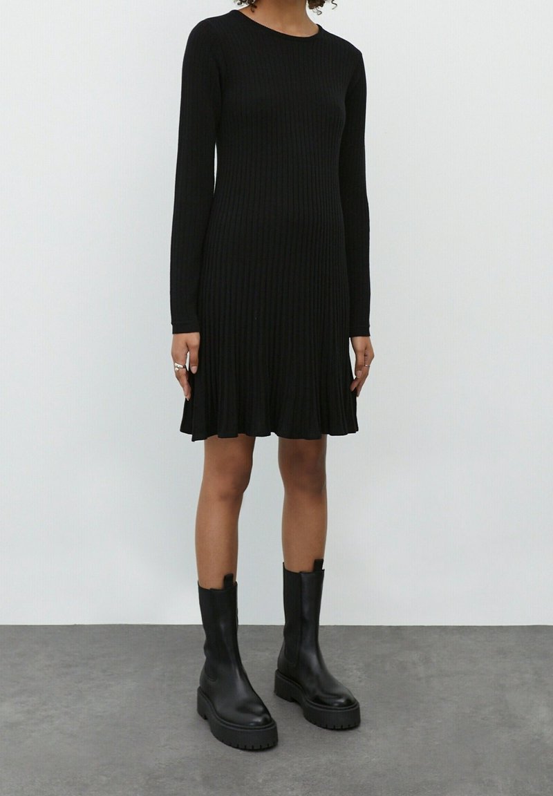 Robe noire à manches longues avec des côtes, dotée d'un ourlet évasé, associée à des bottes noires à semelle épaisse montantes jusqu'au genou. Texture lisse et silhouette ajustée.