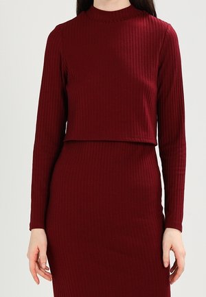 Nainen, joka pukeutunut punertavan tummanpunaisen väriseen, ribbineulottuun pitkähihaiseen crop-toppiin ja samaan sävyyn sopivaan korkeavyötäröiseen hameeseen, yksinkertaisen vaalean taustan edessä.