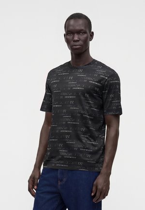 Homme portant une chemise noire à manches courtes avec un motif répétitif du texte « EMPORIO ARMANI » et un jean bleu foncé, debout devant un fond uni.