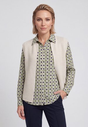 Donna con capelli biondi corti che indossa una camicia verde a fantasia e un gilet lavorato a maglia color crema, posando con una mano in tasca davanti a uno sfondo semplice.