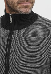 Jersey con cremallera de patrón en blanco y negro, con un alto cuello acanalado y una textura suave. Presenta un detalle de cremallera metálica.