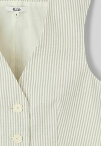 Débardeur sans manches en crème légèrement texturé avec des rayures verticales bleues, doté d'un décolleté en V profond et de trois boutons beiges sur le devant.