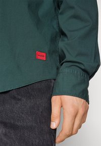 Main droite d'une personne portant une chemise à manches longues vert foncé avec un écusson rouge "HUGO" et un pantalon en jean noir.