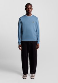 Felpa azzurra con colletto rotondo, polsini a coste e un piccolo logo giallo. Indossata con pantaloni neri a gamba larga e sneakers grige.