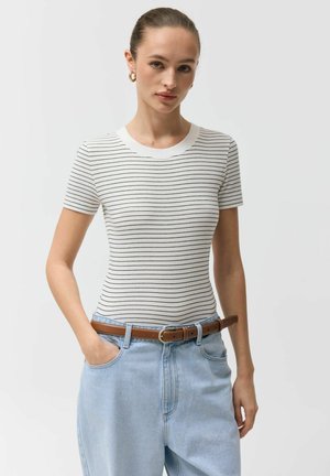 Kobieta z zaczesanymi do tyłu włosami, ubrana w białą prążkowaną koszulkę z krótkim rękawem z cienkimi czarnymi paskami, jasnoniebieskie dżinsy i brązowy pasek.