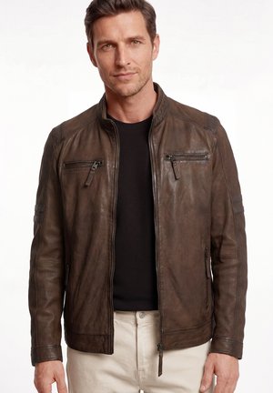 Mann trägt eine dunkelbraune Lederjacke über einem schwarzen Hemd und hellfarbigen Hosen, steht vor einem schlichten weißen Hintergrund.