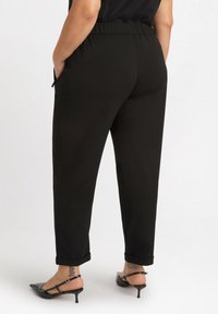 Czarne, tapered spodnie z elastycznym pasem, bocznymi kieszeniami i podwiniętymi mankietami, w zestawieniu z czarnymi sandałami na obcasie i widocznymi tatuażami na kostkach.