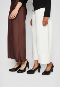 Chaussures noires à talons hauts avec des orteils ronds, associées à une jupe en satin marron et un pantalon large blanc, mettant en avant des matériaux lisses et des lignes épurées.