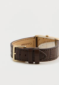 Καφέ δερμάτινο λουρί ρολογιού με ορθογώνια χρυσή θήκη, που διαθέτει υφή και κούμπωμα. Το λογότυπο είναι ορατό από το εσωτερικό.