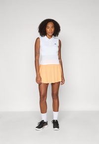 Vit ärmlös polo med krage, kombinerad med en orange veckad kjol. Svarta träningsskor och vita strumpor fulländar outfiten.
