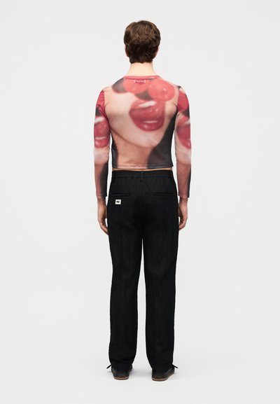 Persona con pantalones negros y una camiseta de manga larga ajustada con un gran estampado de labios rojos y cerezas en la espalda, de pie de espaldas.