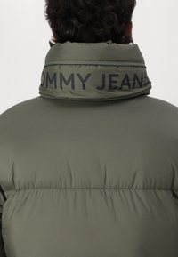 Chaqueta acolchada en color verde oliva, con un alto cuello que lleva la inscripción "TOMMY JEANS", textura suave y cierre de cremallera en el cuello.