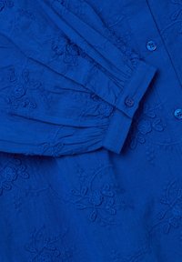 Camicia blu ricamata con maniche lunghe, presenta motivi floreali, chiusura a bottone e pieghe lungo la manica per un tocco di texture in più.