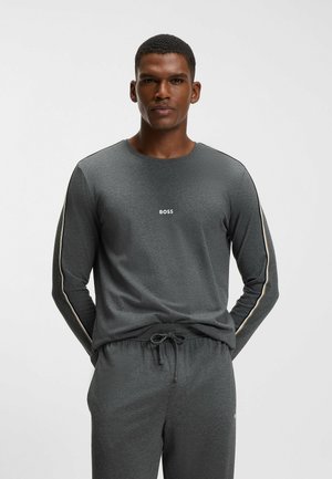 Man draagt een donkergrijs lang mouwen shirt en bijpassende broek met BOSS-logo op de borst en strepen op de mouwen, staand met handen achter de rug.