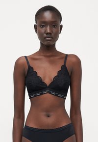 Bralette in pizzo nero con bordi ondulati, spalline sottili e una banda elastica con logo. Il design presenta una scollatura a V profonda e una texture morbida.