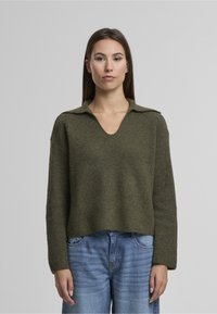 Sweter z dzianiny w oliwkowo-zielonym kolorze, z dekoltem w serek i kołnierzem, z długimi rękawami i luźnym krojem, noszony na jasno-niebieskich dżinsach.