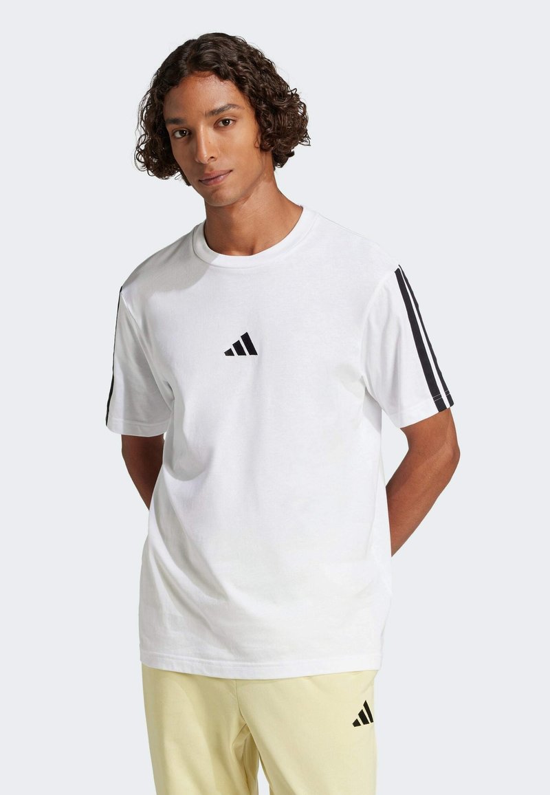 Biela bavlnená tričko s čiernym logom Adidas na hrudi a čiernymi pruhmi na rukávoch. Okrúhly výstrih a voľný strih.