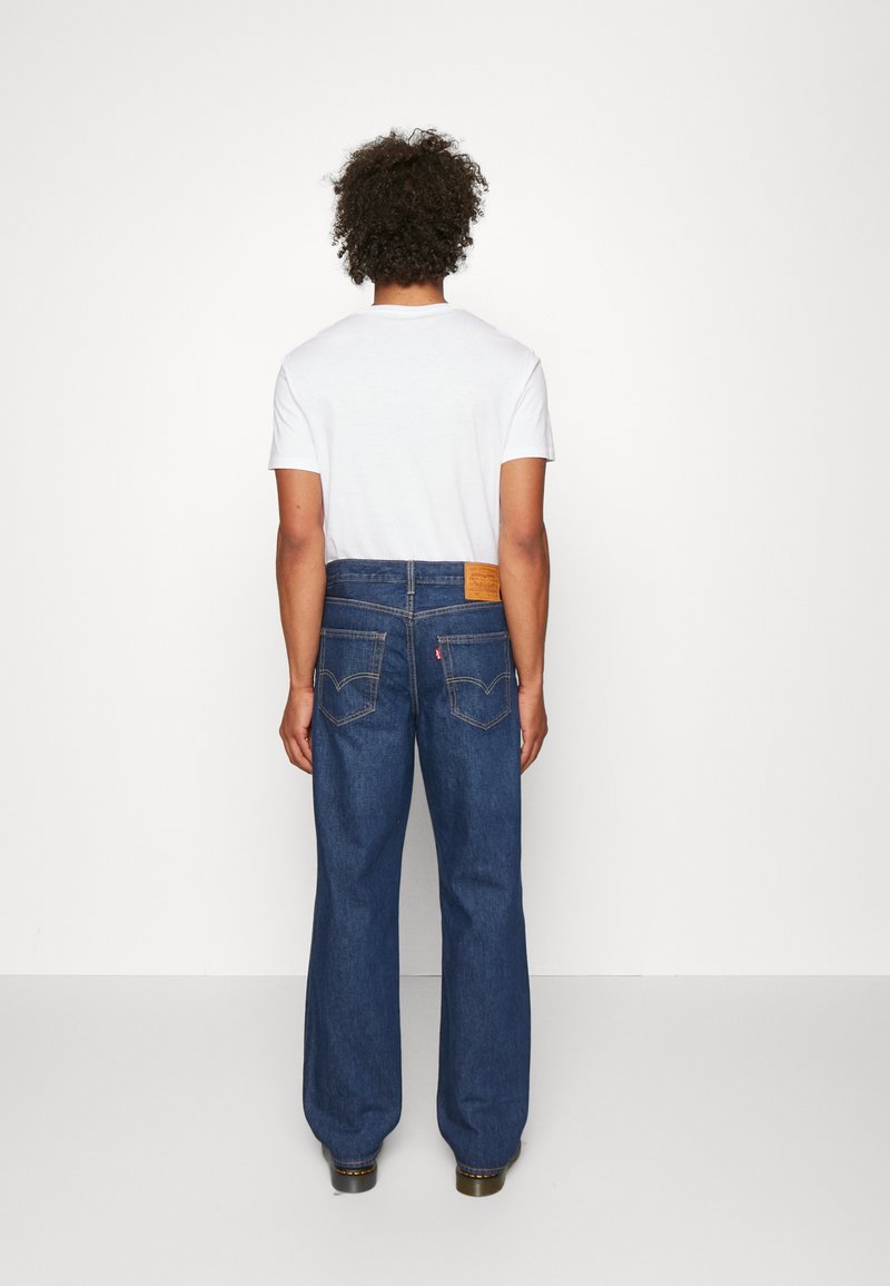 Una persona che indossa una t-shirt bianca e jeans di denim blu, con una toppa in pelle in vita e due tasche posteriori con dettagli di cucitura.