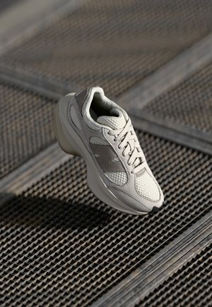 Baskets gris clair avec panneaux en mesh et suède, flottant au-dessus d'une surface en grille métallique texturée à la lumière naturelle.