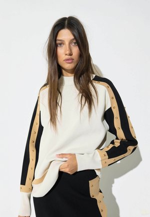Mujer con pelo largo y castaño, vistiendo un suéter blanco con rayas tachonadas negras y beige en las mangas y una falda a juego, de pie frente a un fondo blanco.