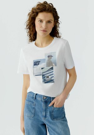 Junge Frau mit lockigem Haar, die ein weißes T-Shirt mit Fotomotiven und eine blaue, hochtaillierte Jeans trägt, eine Hand in der Tasche, neutraler Hintergrund.