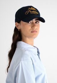 Musta lippalakki, jossa on keltainen brodeerattu teksti "Kielletty 10119 Berliini", kaarevalla lieriöllä ja sileällä kankaalla. Yhdistetty vaaleansiniseen kauluspaitaan.