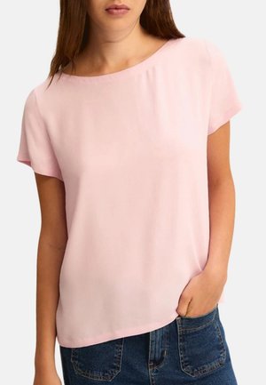 Femme portant un chemisier rose clair à manches courtes avec un col bateau et un jean en denim bleu foncé, main dans la poche.