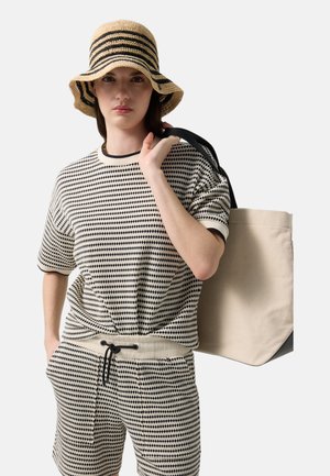 Woman wearing black and white patterned shirt and shorts, carrying beige tote bag, and wearing beige hat with black stripes.