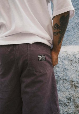 Osoba w ciemnofioletowych szortach z naszywką "Limited Code" na tylnej kieszeni i białej koszuli, stojąca na tle fakturowanej jasnoniebieskiej ściany.