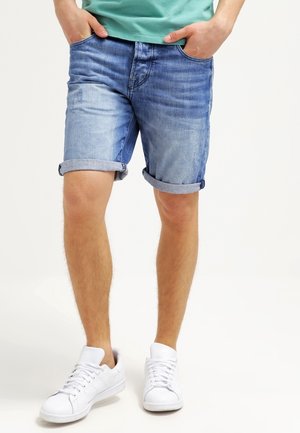 Jasnoniebieskie szorty dżinsowe z efektem sprania, podwiniętymi mankietami i pięcioma kieszeniami, noszone z białymi trampkami i zieloną koszulką.