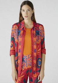 Rood en oranje gestreept shirt met bloemendessign, knoopsluiting aan de voorkant en opgerolde mouwen, gecombineerd met een aansluitend oranje topje, gedragen met bijpassende broek.