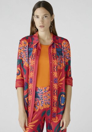 Rood en oranje gestreept shirt met bloemendessign, knoopsluiting aan de voorkant en opgerolde mouwen, gecombineerd met een aansluitend oranje topje, gedragen met bijpassende broek.