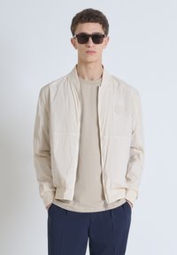 Beige bomberjack met een rits aan de voorkant, ribgebreide manchetten en een logopatch. Draagt over een lichtgrijs t-shirt en gecombineerd met marineblauwe broeken.