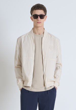 Chaqueta bomber beige con cremallera en la parte delantera, puños acanalados y un parche con logo. Llevada sobre una camiseta gris clara y combinada con pantalones azul marino.