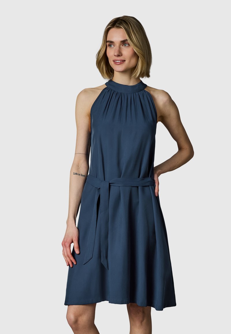 Donna che indossa un abito senza maniche blu scuro con collo halter e cinturino in vita, in piedi con una mano sul fianco, sfondo neutro.