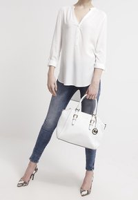 Hvidt håndtaske med sølvhardware, struktureret design og logocharms. Model iført en hvid top og blå jeans med slangeskinds-hæle.