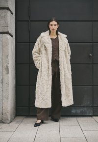 Lang, cremefarvet faux fur frakke med en bred krave og struktureret overflade, båret over et brunt outfit, mod en mørk baggrund.