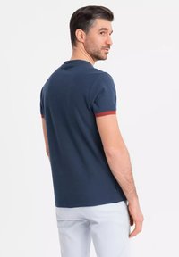 Polo de color azul marino con mangas cortas, ribete rojo en el cuello y las mangas, y una textura suave. Combinado con pantalones de color claro.