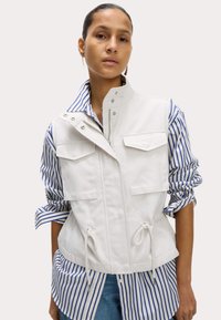 Gilet blanc avec un col haut, des poches latérales et un ourlet à cordon, superposé sur une chemise rayée bleu et blanche. Texture de tissu lisse.