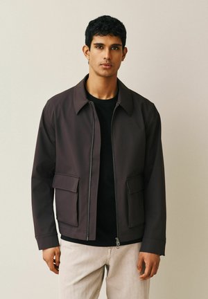 Man standing wearing a dark brown zip-up jacket with large front pockets, black shirt, and light beige pants against a plain background.