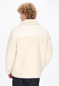 Hellbeige Fleecejacke mit einem strukturierten, flauschigen Körper und einem glatten, hellen Oberteil; verfügt über einen hohen Kragen und einen entspannten Schnitt.