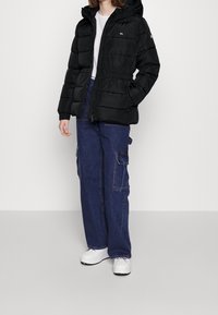 Giacca piumino nera con cappuccio, vita elastica e chiusura con zip, abbinata a pantaloni cargo in denim scuro e scarpe da ginnastica bianche.