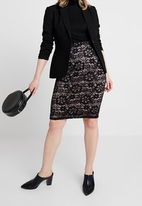 Nainen, joka on pukeutunut mustaan bleiseriin, kukkakuvioiseen pitsihameeseen, mustiin korokepohjaisiin kengännauhoihin, pitäen pyöreää mustaa käsilaukkua yksinkertaista taustaa vasten.