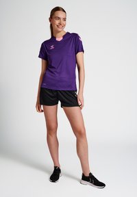 Purple athletic t-shirt with pink shoulder accents, paired with black shorts. Model stands, wearing black athletic shoes, on a neutral background.