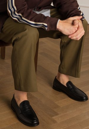 Zwarte leren instapschoenen met een gladde afwerking en gestikte accenten, gecombineerd met olijfgroene broeken, zittend op een houten vloer.